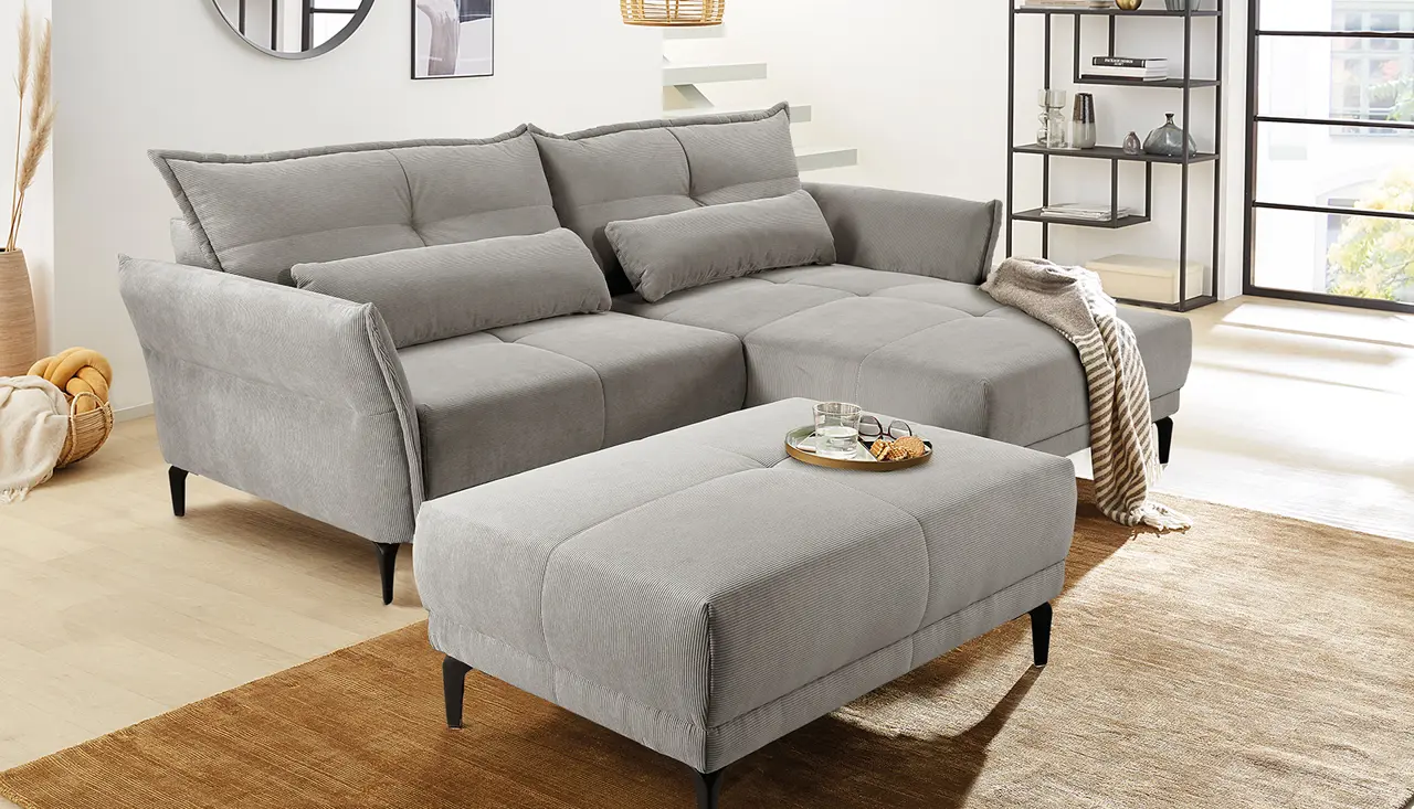 Moderne graue Wohnlandschaft mit großem Hocker und gemütlichen Kissen in hell eingerichtetem Wohnzimmer. Polsterecke mit viel Liegefläche, ideal zum Entspannen. Stilvolle Deko und warmes Tageslicht schaffen ein einladendes Ambiente.